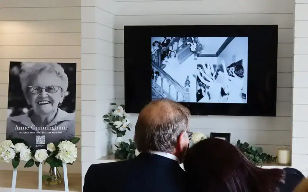 Man and woman embrace in front of a memorial. To the left, a portrait of an elderly lady. A black-and-white photo on the monitor shows memories. - honorpix People viewing a memorial slideshow on a large screen at a funeral service.