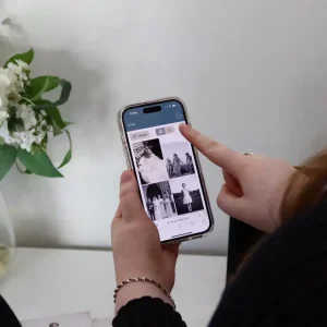 Two women look at photos on a smartphone, pointing to photos of a loved one in a GUESTPIX memorial gallery. A vase of white flowers is in the background, creating a calm and intimate setting.